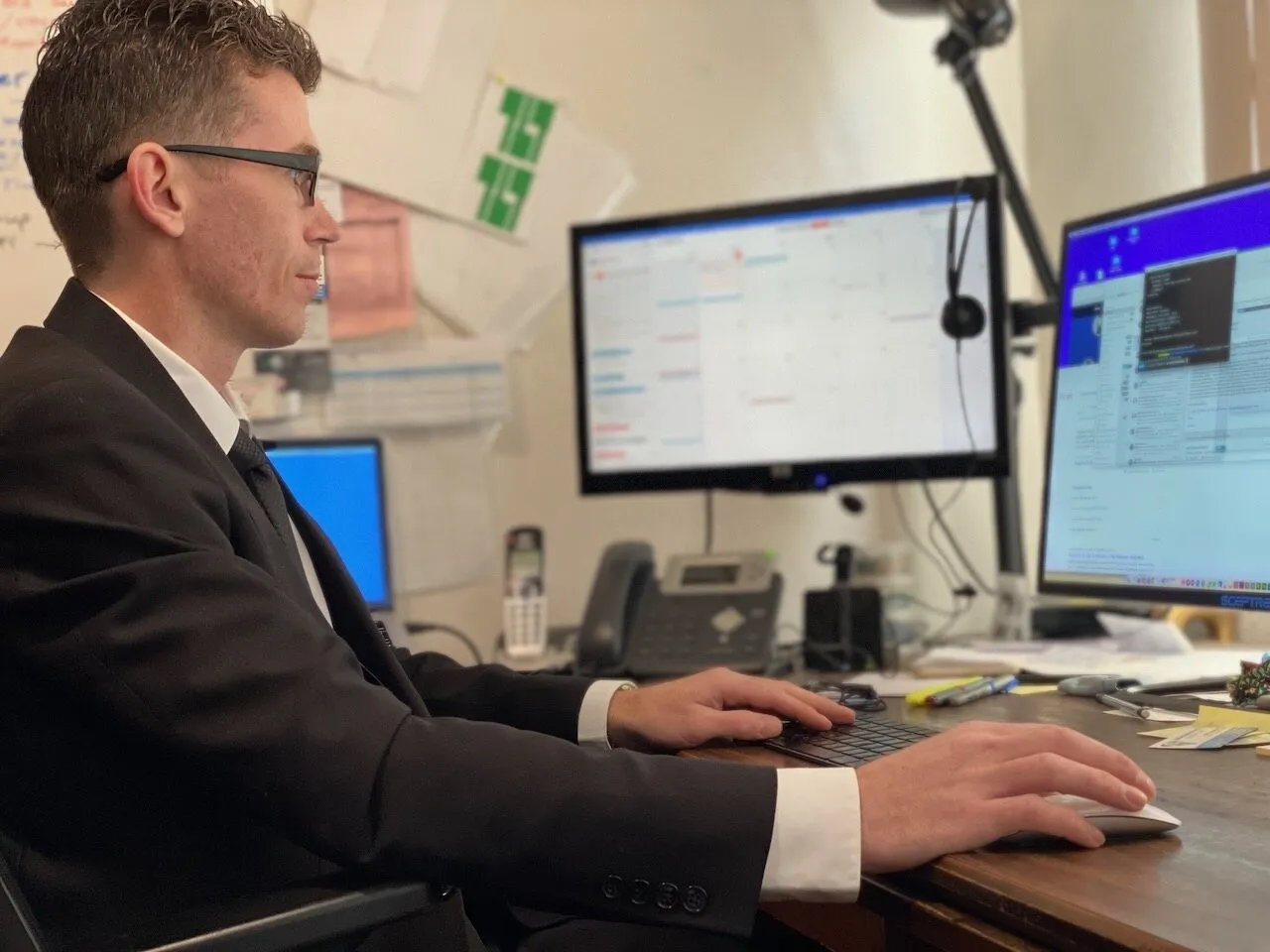 Josh Madrone in a dark suit at his desk, typing, with a calendar on one monitor and a remote session on another, a desk phone and headset beside him