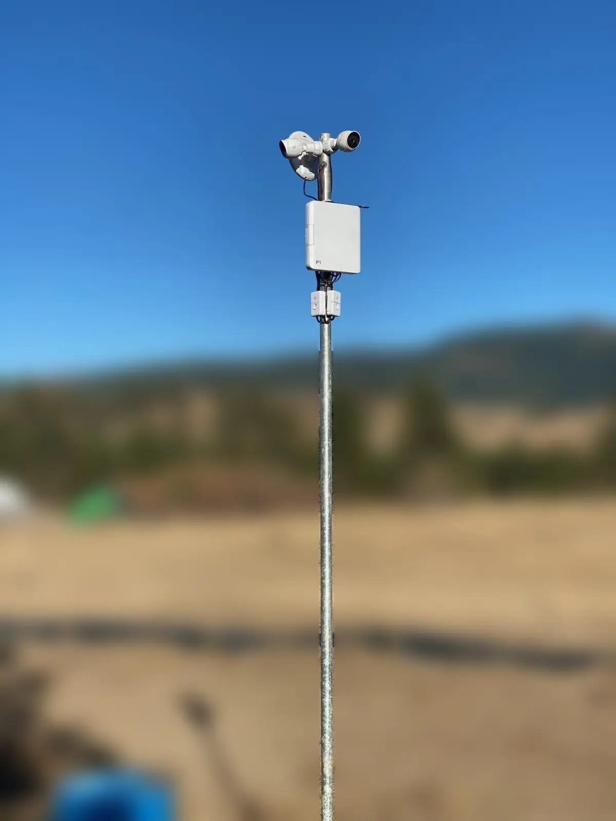 Security cameras on a steel pole with a wireless backhaul radio, sharp against a blurred rural hillside