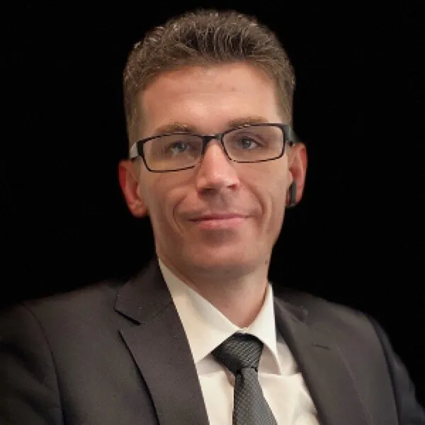 Head-and-shoulders portrait of Josh Madrone in glasses, a dark suit and tie, against a black background