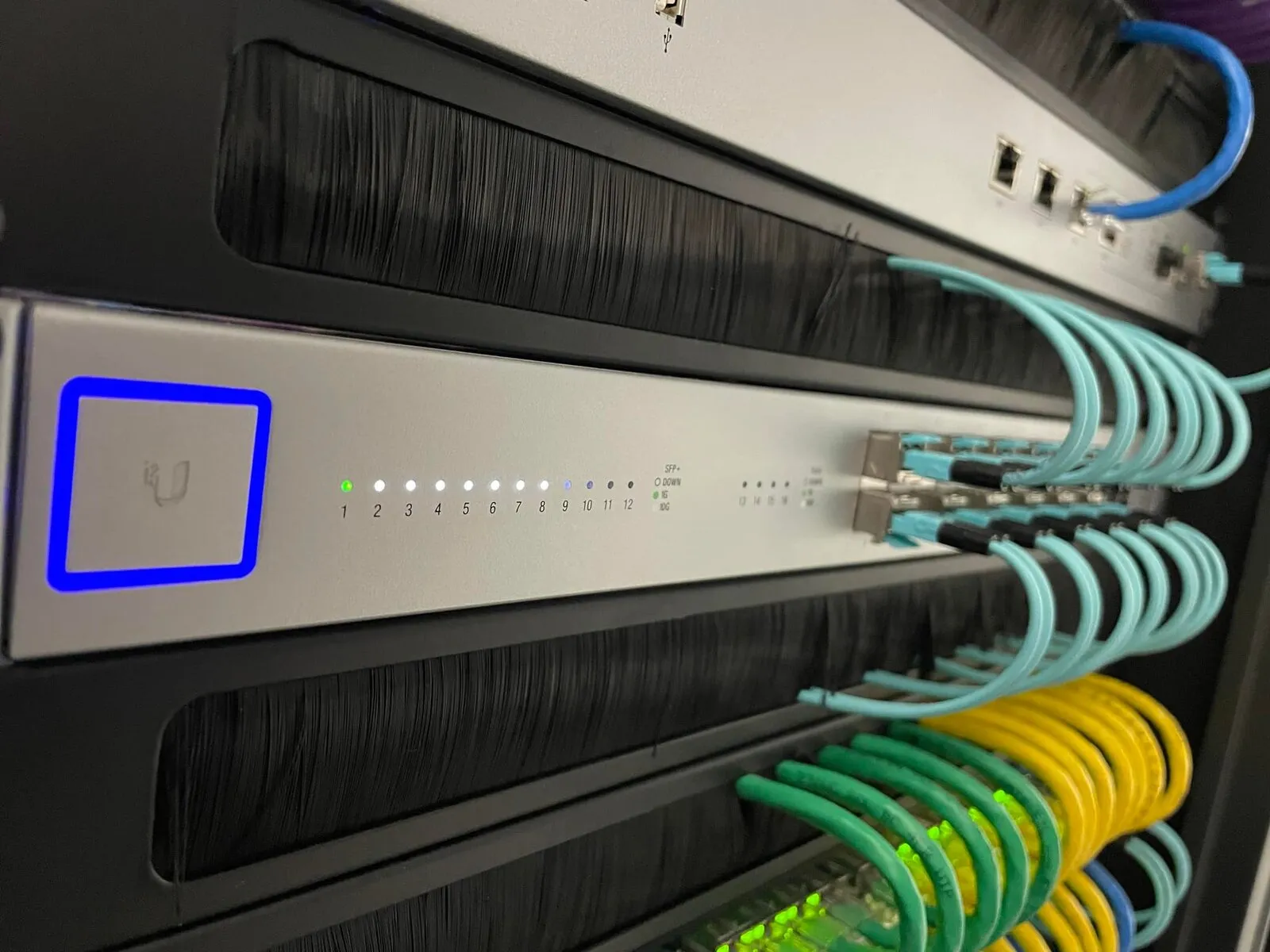 UniFi switch in a rack with neatly routed teal, green and yellow patch cables behind brush-strip panels