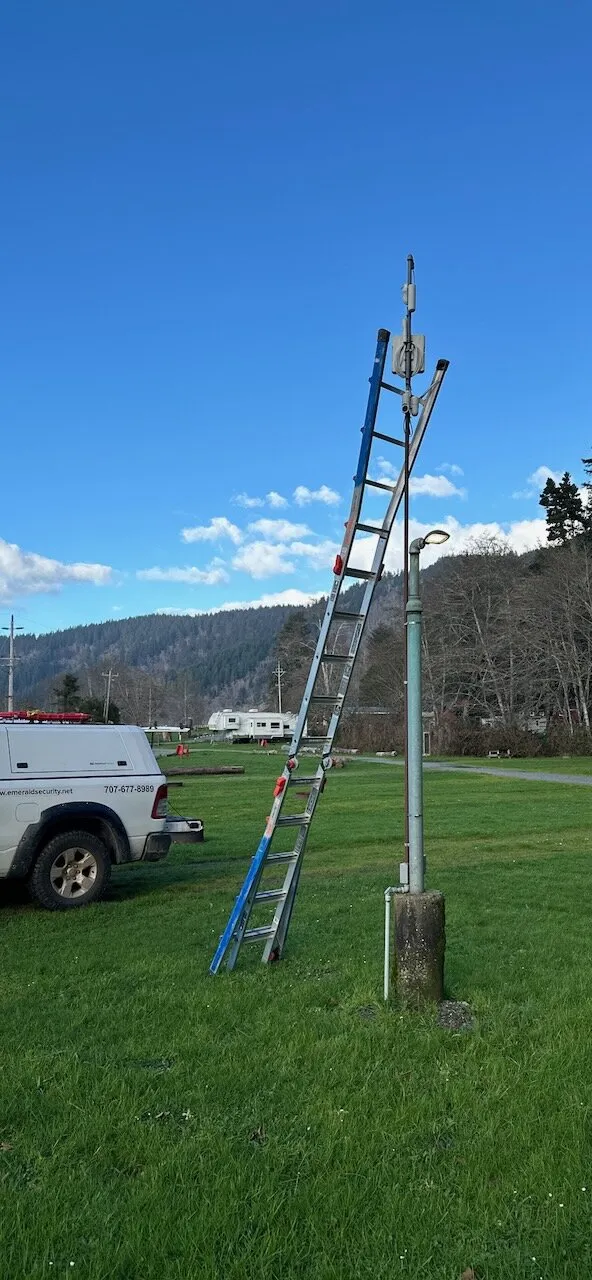 Ladder against a pole carrying a wireless radio and cameras on a grassy site, with an Emerald Security truck parked nearby