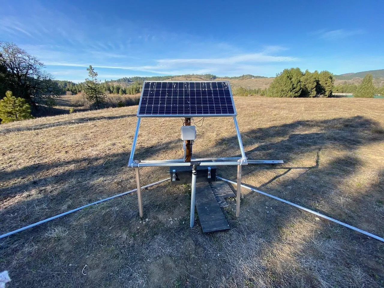 Solar panel on an aluminum ground frame above a battery box, with conduit running out across a dry field