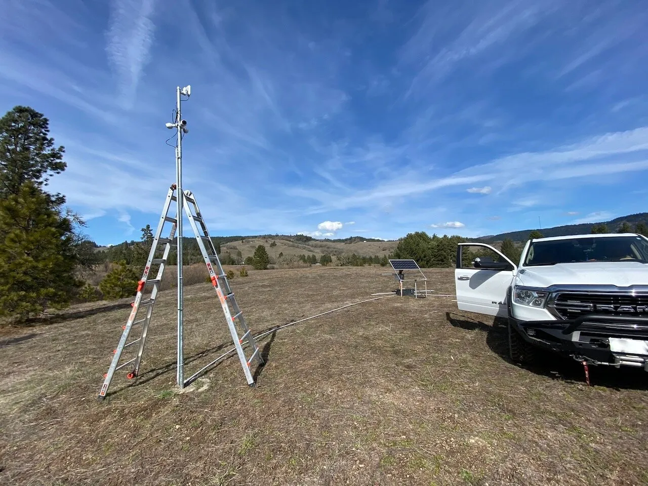 Camera pole with three cameras beside a stepladder, a ground-mounted solar panel and a pickup truck in an open field