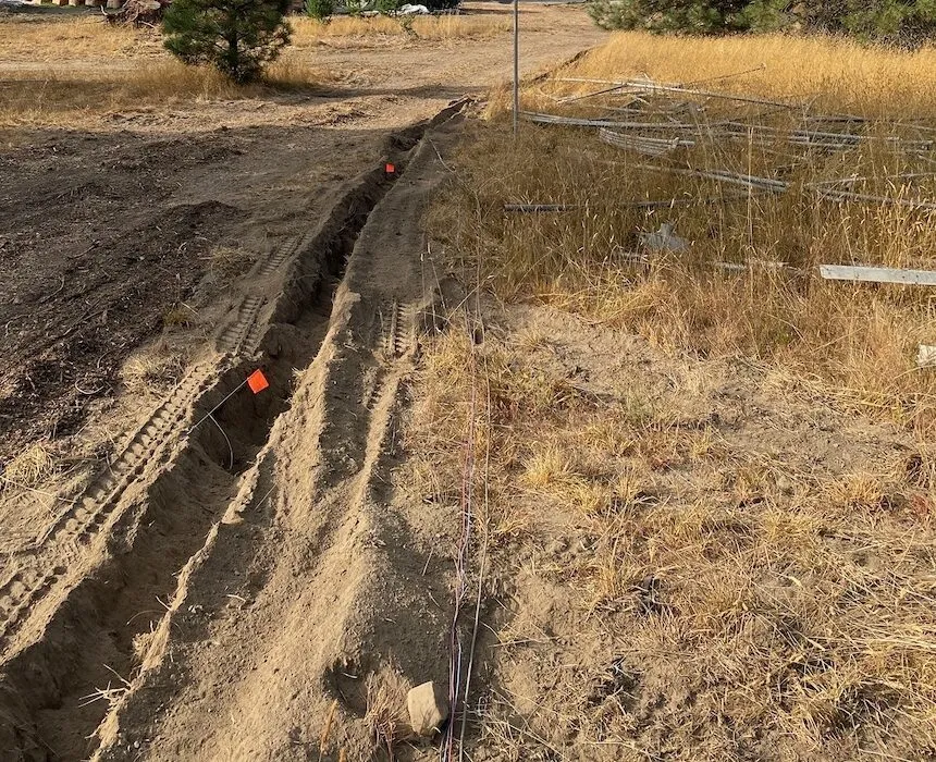 A freshly dug trench across dry, open ground beside a dirt track, marked with orange flags.